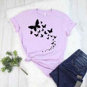 May include: A light purple t-shirt featuring a black butterfly design. The design shows a large butterfly with smaller butterflies trailing behind it. The shirt is paired with blue jeans.