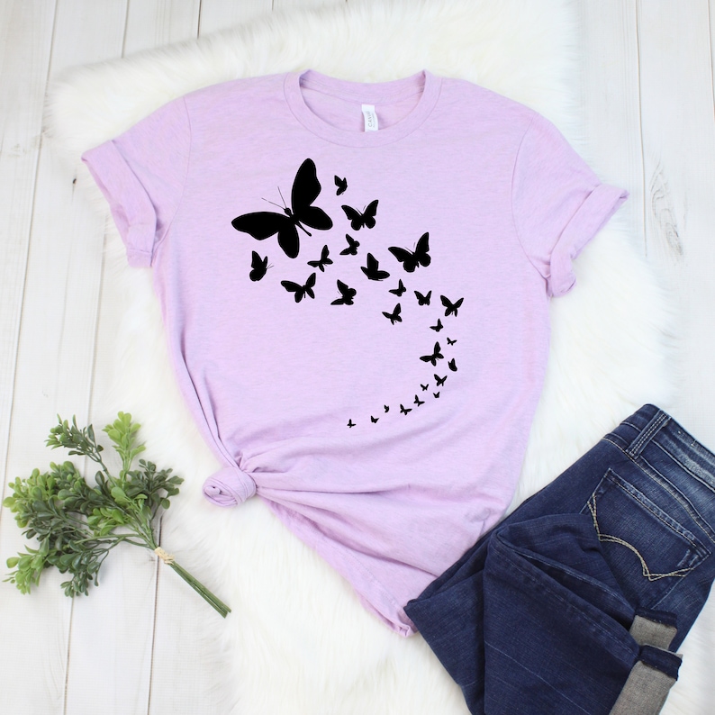 May include: A light purple t-shirt featuring a black butterfly design. The design shows a large butterfly with smaller butterflies trailing behind it. The shirt is paired with blue jeans.