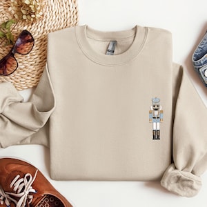 May include: A beige sweatshirt featuring a small embroidered image of a nutcracker in a blue and gold uniform. The nutcracker is standing with his arms by his side.