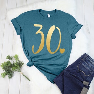 May include: Teal t-shirt featuring the number "30" in gold script and a small gold heart. The shirt is styled with dark blue jeans. The t-shirt is knotted at the waist and is displayed on a white fur-like surface.