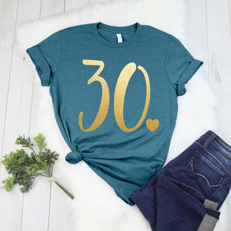 May include: Teal t-shirt featuring the number "30" in gold script and a small gold heart. The shirt is styled with dark blue jeans. The t-shirt is knotted at the waist and is displayed on a white fur-like surface.
