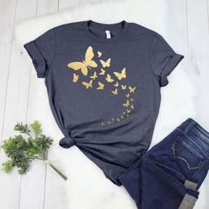 May include: Dark gray t-shirt featuring a gold butterfly design. The design includes a large butterfly with smaller butterflies trailing behind it. The shirt is paired with dark wash denim jeans.