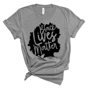 May include: A gray t-shirt with the words "Black Lives Matter" in black script and a silhouette of a person's head. The shirt is knotted at the bottom and has rolled-up sleeves. The shirt is made of a soft material.
