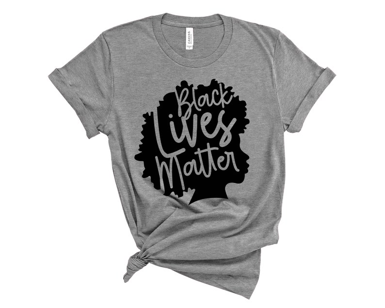 May include: A gray t-shirt with the words "Black Lives Matter" in black script and a silhouette of a person's head. The shirt is knotted at the bottom and has rolled-up sleeves. The shirt is made of a soft material.