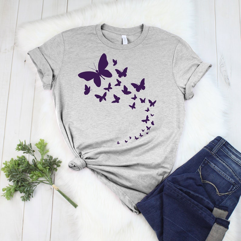 May include: A light gray t-shirt featuring a design of purple butterflies in various sizes, arranged in a flowing pattern. The butterflies appear to be in flight. The shirt is paired with dark blue jeans.