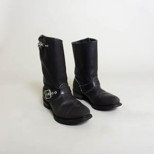 Könnte beinhalten: Ein Paar schwarze Leder-Motorrad-Stiefel mit silbernen Schnallen. Die Stiefel haben eine spitze Zehenpartie und einen niedrigen Absatz.