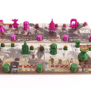 Op de afbeelding: Een spelbord met groene en roze plastic spelstukken. Het bord heeft een bruine achtergrond met illustraties van gebouwen en mensen. De spelstukken zijn gevormd als gebouwen, windmolens en andere objecten.