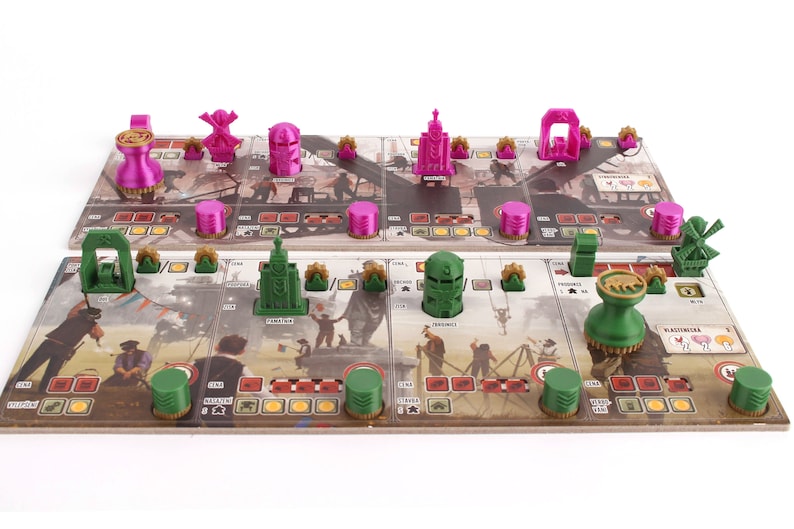 Op de afbeelding: Een spelbord met groene en roze plastic spelstukken. Het bord heeft een bruine achtergrond met illustraties van gebouwen en mensen. De spelstukken zijn gevormd als gebouwen, windmolens en andere objecten.
