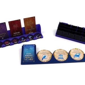Op de afbeelding: Een set paarse en zwarte spelorganizers met kaarten en tokens. De kaarten hebben tekst en illustraties. De tokens hebben illustraties en tekst. De organizers zijn ontworpen om spelcomponenten te bevatten.