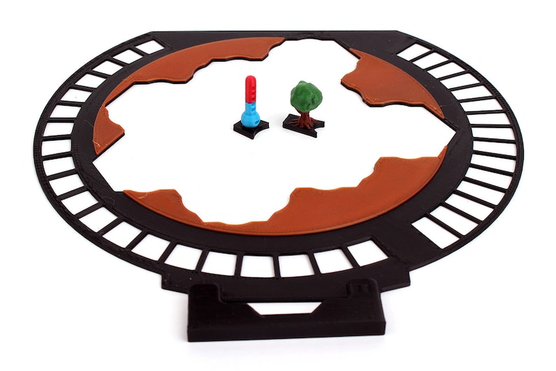 May include: A black circular game board with a brown terrain and a red and blue tower and a green tree on the board.