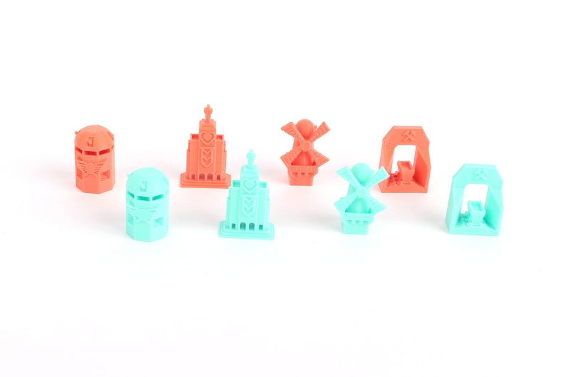 Op de afbeelding: Een set van acht miniatuur 3D-geprinte gebouwen in koraal- en turquoisekleuren. De gebouwen omvatten een windmolen, een kerk, een toren en een poort.