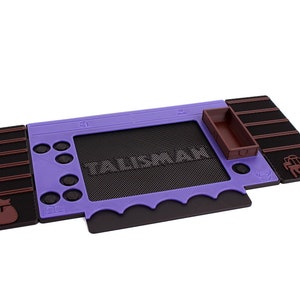 Pu&ograve; includere: Un tappetino da gioco in gomma viola e marrone con la parola "Talisman" stampata su di esso. Il tappetino ha un piccolo vassoio marrone e una piccola figura marrone di un asino.