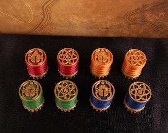 May include: Eight small, round, decorative game pieces with gold trim. Each piece has a different design on the top: a scarab beetle, an atom symbol, and a stylized flower. The pieces are arranged in two rows of four, with the scarab beetle pieces on the ends of each row. The pieces are on a black surface.
