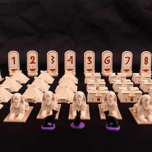 Pode incluir: Um conjunto de jogo de tabuleiro com peças bege e marrom, incluindo pirâmides, obeliscos, estátuas de esfinges e peças numeradas. Há também três figuras de gatos pretos em bases roxas.