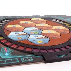 May include: A black and brown board game with a circular, rotating section in the center. The center section has a red, brown, and blue hexagonal tile pattern. The board has a black and red temperature gauge and a black and green tree.