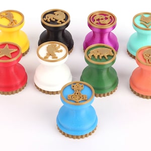 Op de afbeelding: Negen kleurrijke, ronde, dubbelzijdige stempels met een gouden rand. Elke stempel heeft een ander symbool, waaronder een ster, een paard, een leeuw, een wolf, een vis, een everzwijn, een schedel en een hamer.