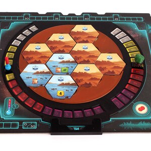 May include: A black and blue game board with a circular, rotating section in the center. The center section features a hexagonal grid of colorful tiles, a red and blue temperature gauge, and a red and white thermometer. The board also includes a black and white timer, a red and white button, and a green and brown tree.
