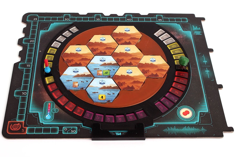 May include: A black and blue game board with a circular, rotating section in the center. The center section features a hexagonal grid of colorful tiles, a red and blue temperature gauge, and a red and white thermometer. The board also includes a black and white timer, a red and white button, and a green and brown tree.