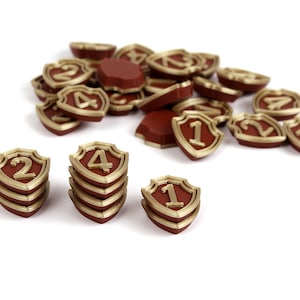 Könnte beinhalten: Ein Satz roter und goldener schildförmiger Spielsteine mit den Zahlen 1, 2 und 4 darauf gedruckt. Die Spielsteine sind in Stapeln von drei gestapelt.