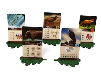Puede incluir: Cinco cartas de juego con ilustraciones de animales y texto en un idioma extranjero. Las cartas se muestran en soportes de hojas verdes.
