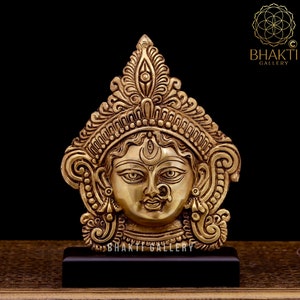 May include: A gold-colored metal sculpture of the Hindu goddess Durga. The sculpture is detailed with intricate carvings and features a third eye on the forehead. The sculpture is displayed on a black wooden base.