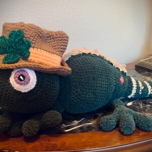 May include: A handmade, dark green crochet lizard wearing a brown hat with a green shamrock. The lizard has a large, multi-colored eye, an orange tongue, and green feet. The tail is striped with white and green.
