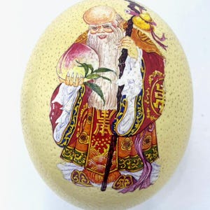 May include: A hand-painted decorative ostrich egg with a detailed illustration of a smiling, bearded figure in ornate robes. The figure holds a peach and staff, set against a yellow background.
