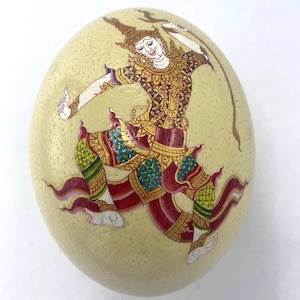 Könnte beinhalten: Ein eierschalenfarbenes Ei ist mit einer detaillierten Malerei eines traditionellen thailändischen Tänzers verziert. Der Tänzer trägt goldene, rote und türkisfarbene Kleidung und hält einen Bogen. Das Kunstwerk ist auf dem Ei zentriert und zeigt das komplizierte Design.