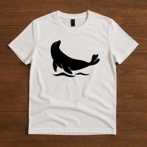 May include: White t-shirt featuring a black silhouette of a seal. The seal is depicted in a stylized pose, with its body curved and flippers extended, set against a simple wave design. This graphic tee is suitable for casual wear.