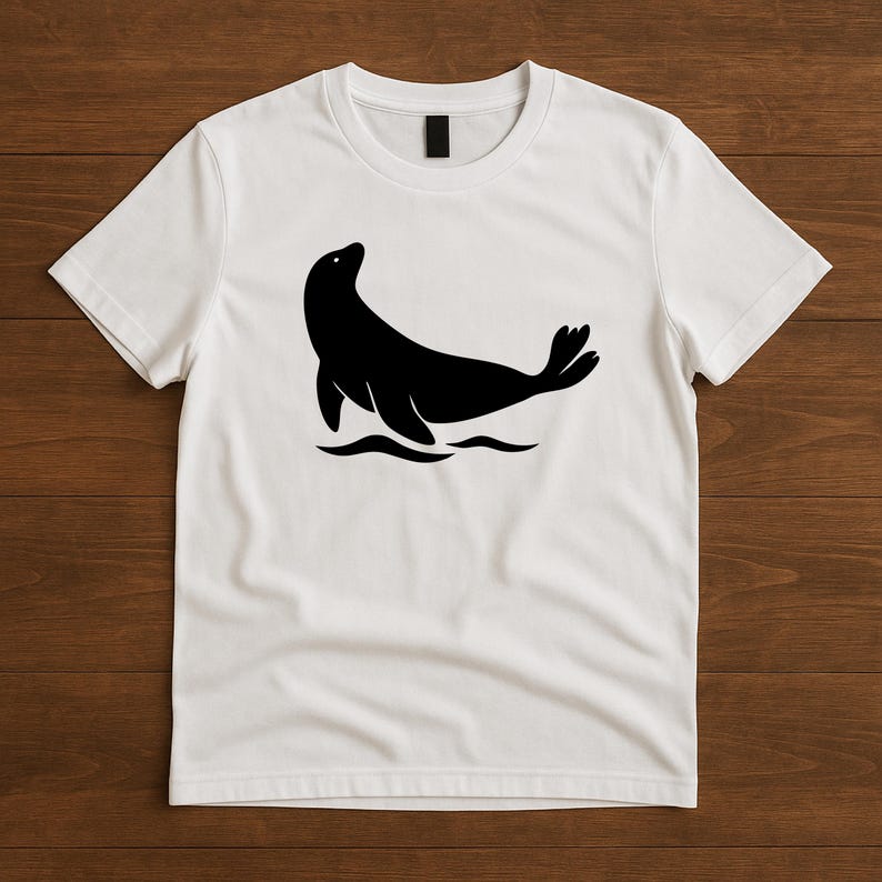 May include: White t-shirt featuring a black silhouette of a seal. The seal is depicted in a stylized pose, with its body curved and flippers extended, set against a simple wave design. This graphic tee is suitable for casual wear.