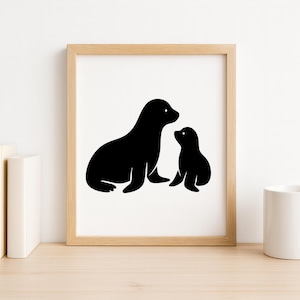May include: A framed print featuring a black silhouette of a mother seal and her pup. The artwork is displayed on a wooden surface, accompanied by books and a white mug.