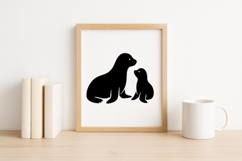 May include: A framed print featuring a black silhouette of a mother seal and her pup. The artwork is displayed on a wooden surface, accompanied by books and a white mug.