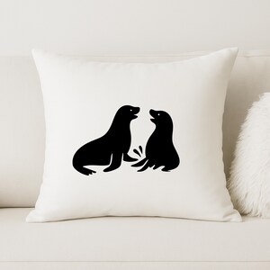 May include: A square, off-white throw pillow featuring a black graphic of two seals facing each other. The seals are stylized with simple shapes and outlines. The pillow is on a white couch.