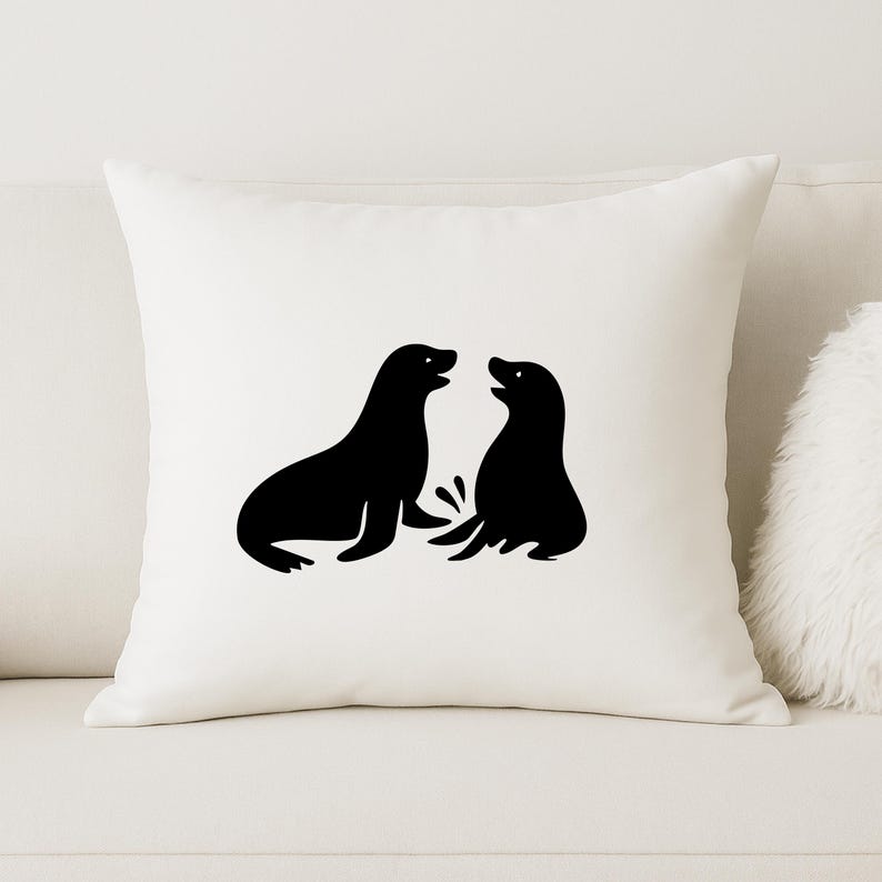 May include: A square, off-white throw pillow featuring a black graphic of two seals facing each other. The seals are stylized with simple shapes and outlines. The pillow is on a white couch.