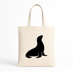 May include: A cream-colored tote bag featuring a black silhouette of a seal. The seal is depicted in a seated position, looking upwards. The bag has two handles and is made of a canvas-like material, suitable for carrying groceries or personal items.