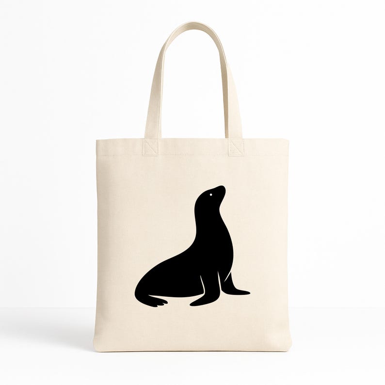 May include: A cream-colored tote bag featuring a black silhouette of a seal. The seal is depicted in a seated position, looking upwards. The bag has two handles and is made of a canvas-like material, suitable for carrying groceries or personal items.