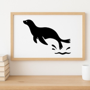 May include: A framed print featuring a black silhouette of a seal leaping through water. The artwork is set against a white background within a light wood frame. The image is displayed on a wooden surface with a stack of books and a white mug.