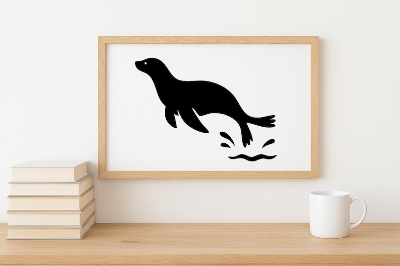 May include: A framed print featuring a black silhouette of a seal leaping through water. The artwork is set against a white background within a light wood frame. The image is displayed on a wooden surface with a stack of books and a white mug.