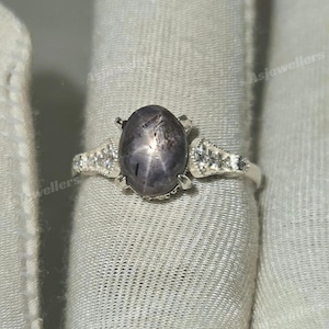 Peut inclure: Bague en argent ornée d'une pierre précieuse ovale gris foncé avec un motif en forme d'étoile. La bague comporte de petites pierres d'accent claires de chaque côté de la pierre principale, serties sur un anneau en argent. La bague est présentée sur un tissu blanc.