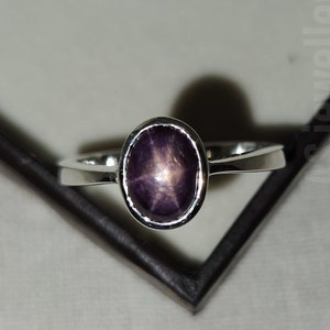May include: A silver ring featuring an oval, star-shaped gemstone with a deep purple hue. The gemstone is set in a simple, polished silver band. The ring is displayed against a neutral background.