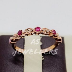 Puede incluir: Un anillo de banda de oro rosa con una fila de pequeñas piedras preciosas rojas y piedras preciosas transparentes más pequeñas engastadas en un diseño delicado. El anillo está en un expositor de terciopelo negro.