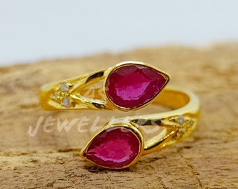 May include: A gold bypass ring featuring two pear-shaped, deep pink rubies at each end. The gold band is accented with small, clear stones near the rubies. The word JEWELLERS is partially visible in white text across the center of the ring.