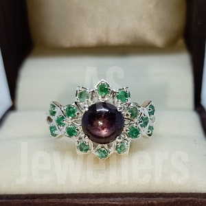 Op de afbeelding: Een zilveren ring met een grote, donkerpaarse edelsteen omringd door kleinere groene edelstenen. De ring ligt in een bruine fluwelen ringdoos.