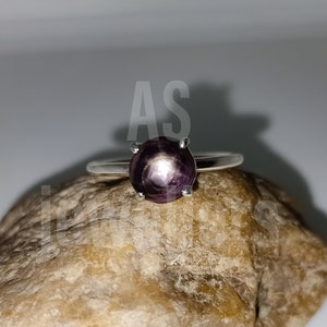 Może przedstawiać: Srebrny pierścionek z ciemnofioletowym kamieniem szlachetnym osadzonym w szponach. Pierścionek leży na brązowym kamieniu.