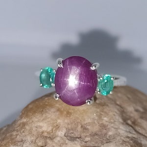 Op de afbeelding: Een zilveren ring met een grote, ronde, roze ster-robijn edelsteen en twee kleinere, ronde, groene edelstenen aan weerszijden.