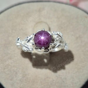 May include: A silver ring featuring a central round cabochon-cut deep purple star gemstone. The band is designed with detailed metallic leaf motifs extending from the setting. The text as jewellers is visible in the background of the image.
