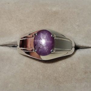 Puede incluir: Un anillo de plata con una gran piedra preciosa ovalada de color púrpura. La piedra preciosa tiene un patrón en forma de estrella en el centro.