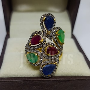 Könnte beinhalten: Ein goldener Ring mit einem birnenförmigen Design, das mit blauen, grünen und roten Edelsteinen besetzt ist. Der Ring ist mit kleinen Diamanten besetzt und hat ein zartes, filigranes Design.