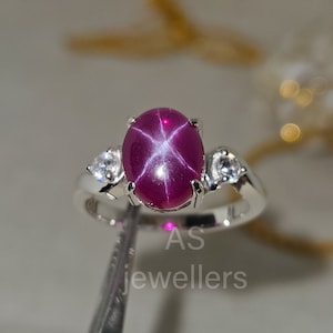 May include: A silver ring with a large, oval-shaped, red star ruby gemstone surrounded by two smaller, round, white gemstones.