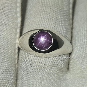 Peut inclure: Une bague en argent avec une grande monture ovale. La monture contient une pierre précieuse ronde, rouge-violet avec un motif en forme d'étoile. La bague est présentée sur une surface blanche.